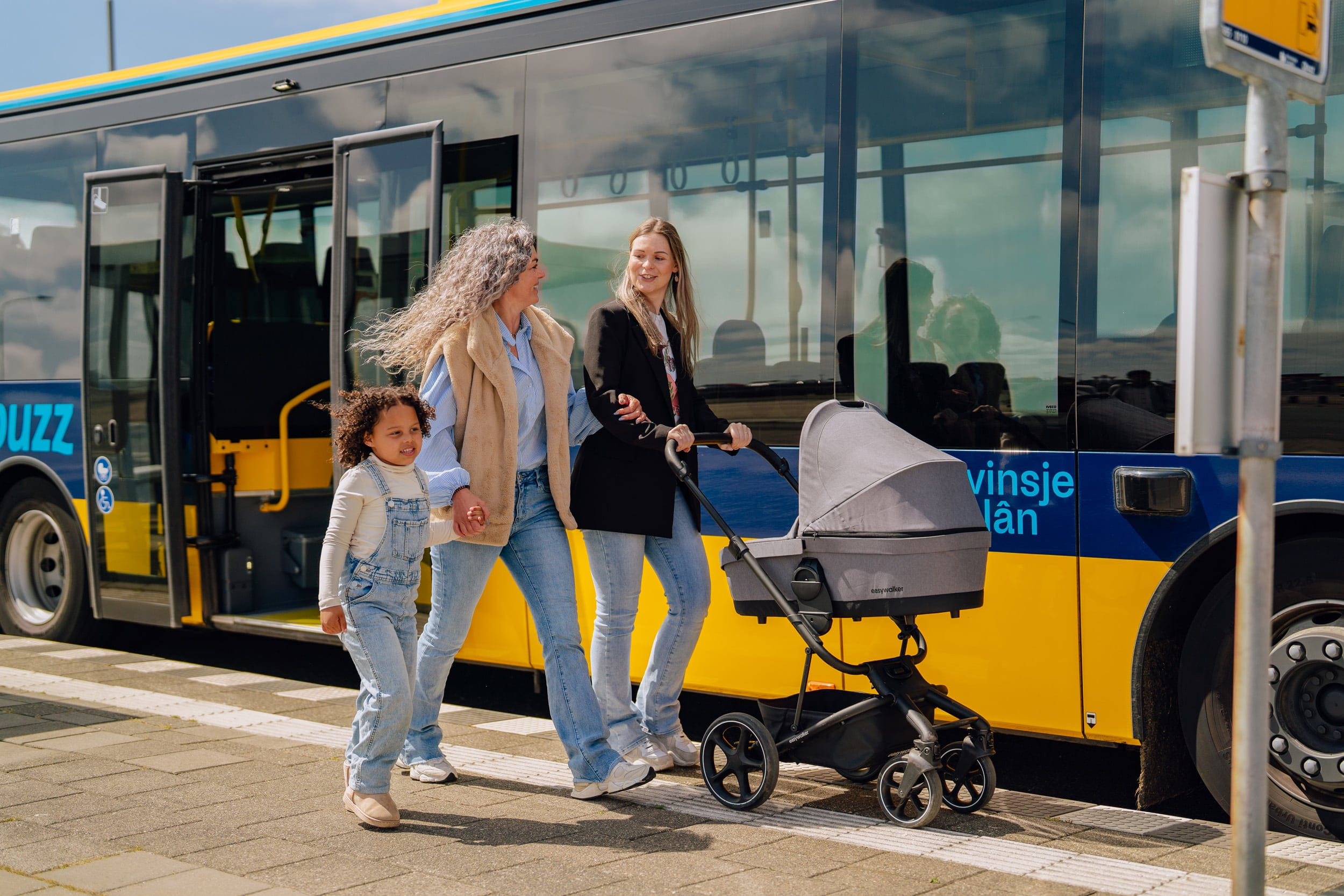 Vriendinnen bij Bushalte met kind en kinderwagen, lachend, wandelend.